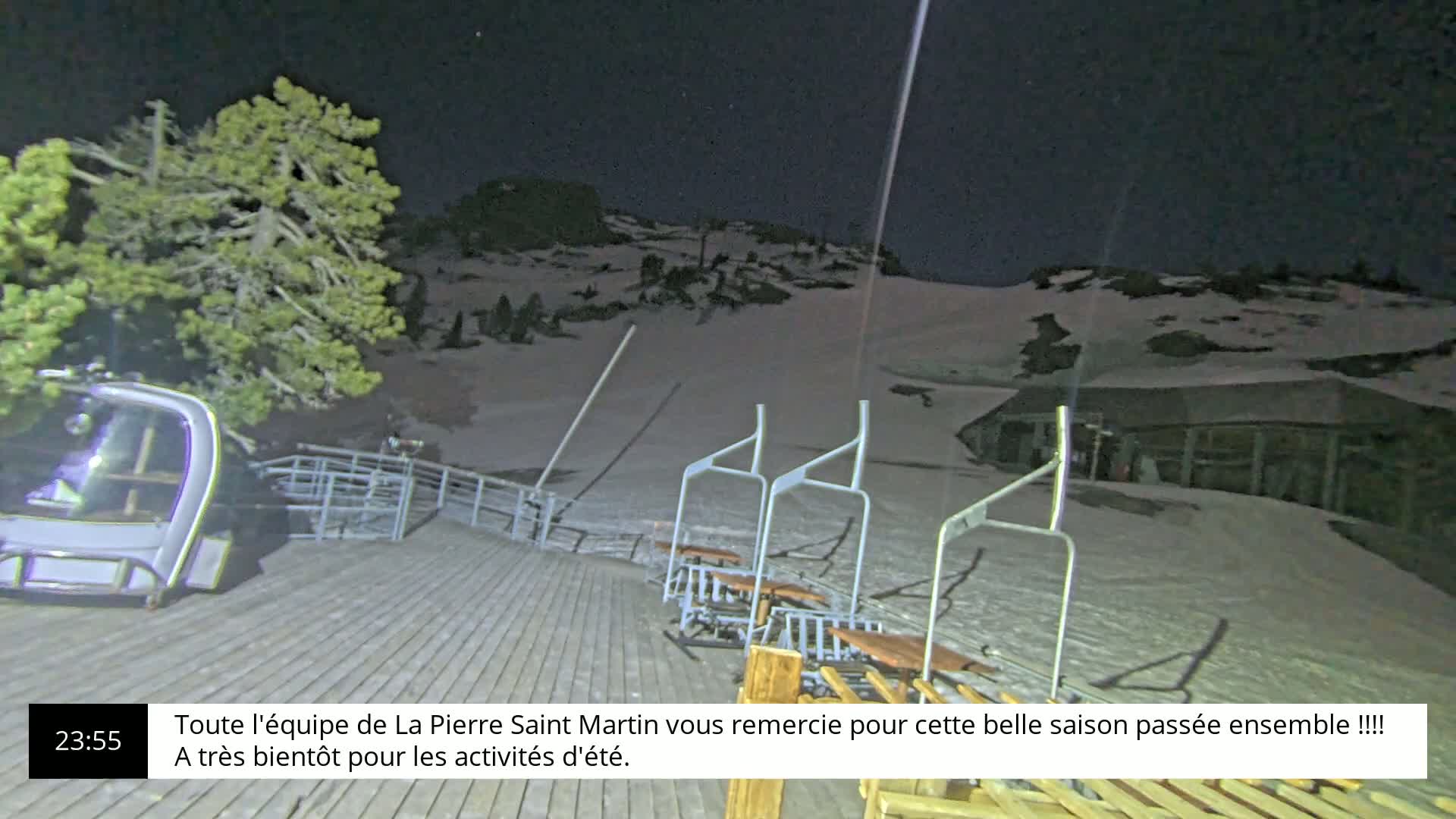 A snow-covered ski slope at night, with several chairlift seats at the bottom and a small building in the background.