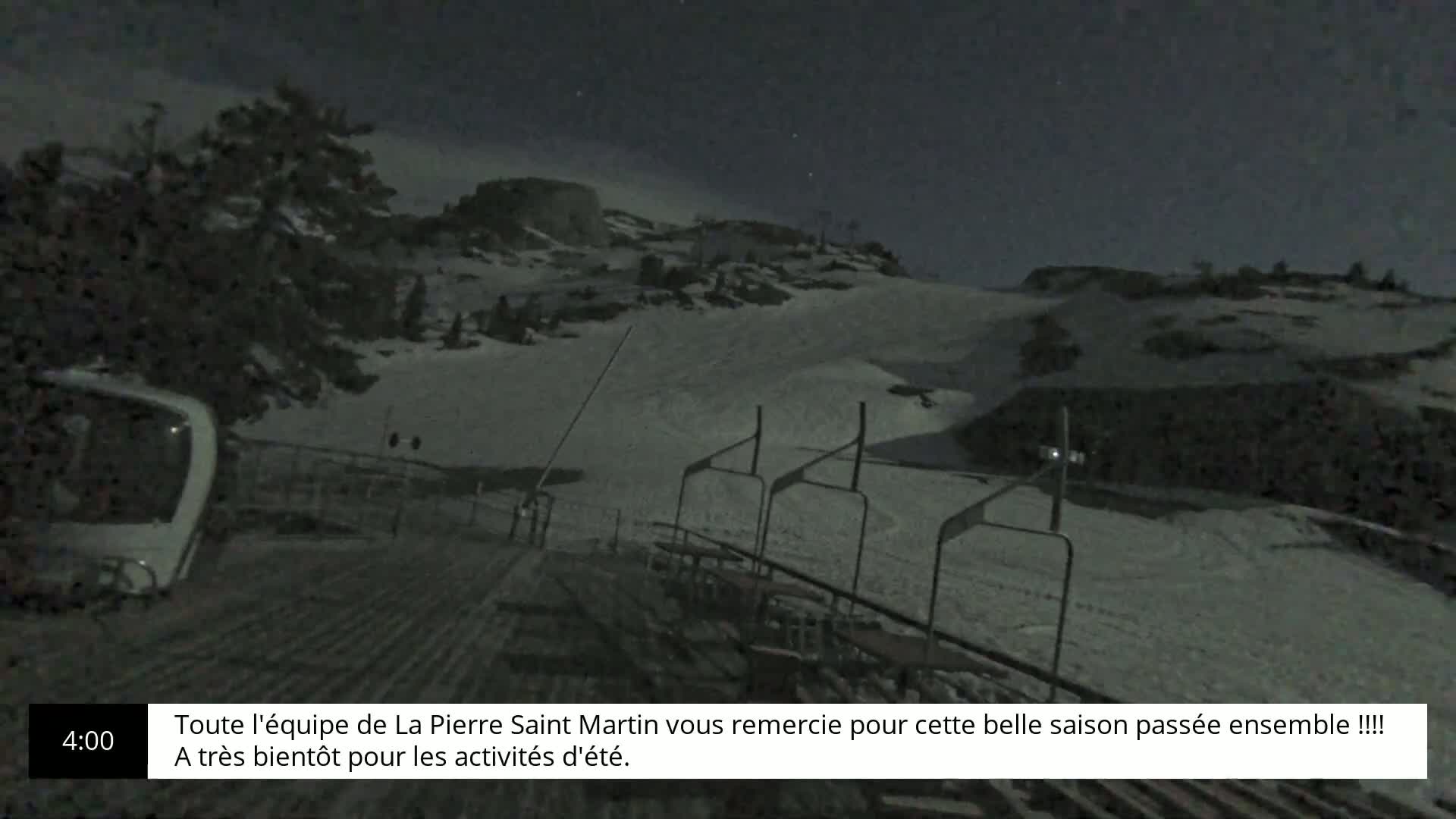 A nighttime view of a snow-covered ski slope with a chairlift and a partially visible ski lift at the bottom, under a dark sky.