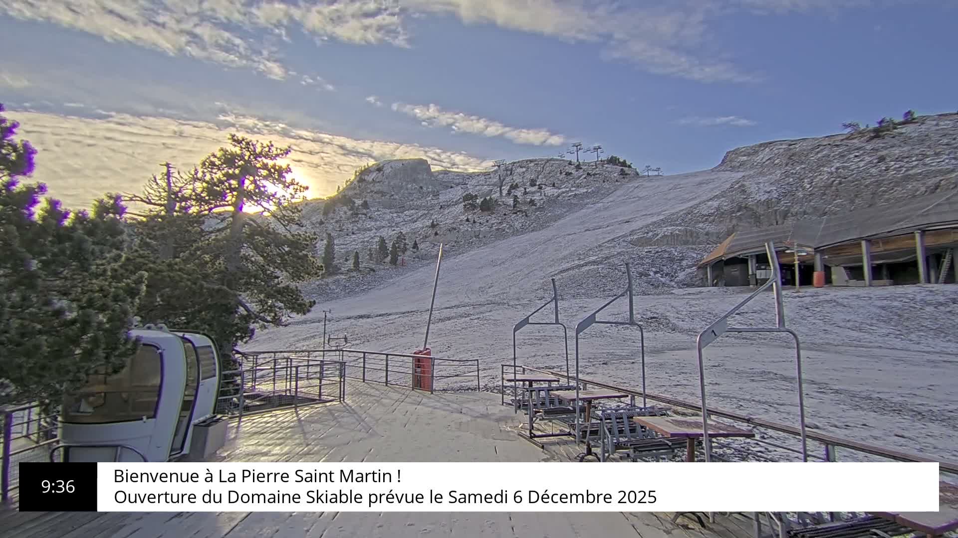 A snow-covered ski slope and mountains are visible under a partly cloudy sky with sunshine, featuring a ski lift gondola on the left and several empty chairlifts in the foreground.