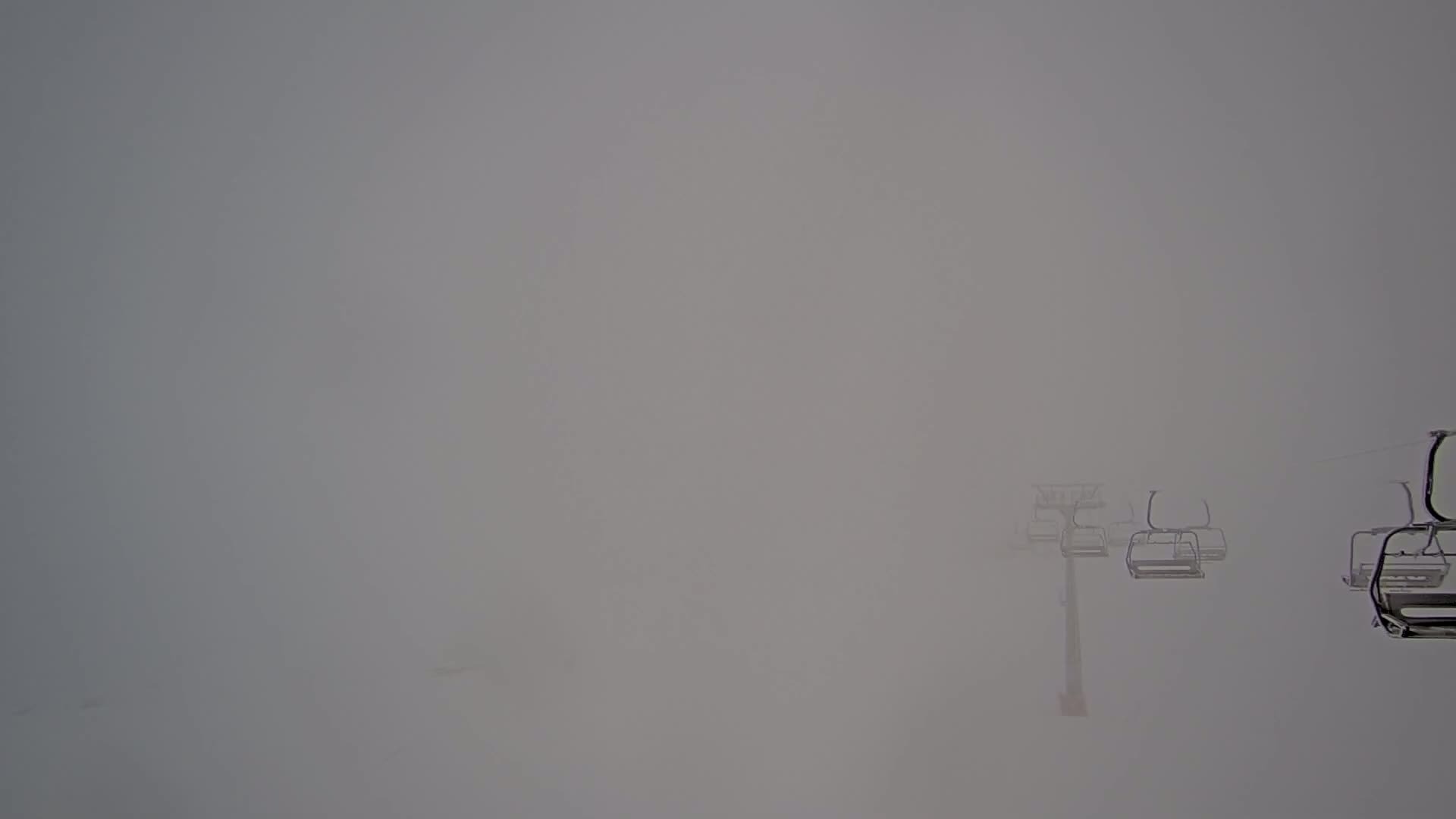 Several empty chairlift cars are visible through a thick fog on a snowy mountain.