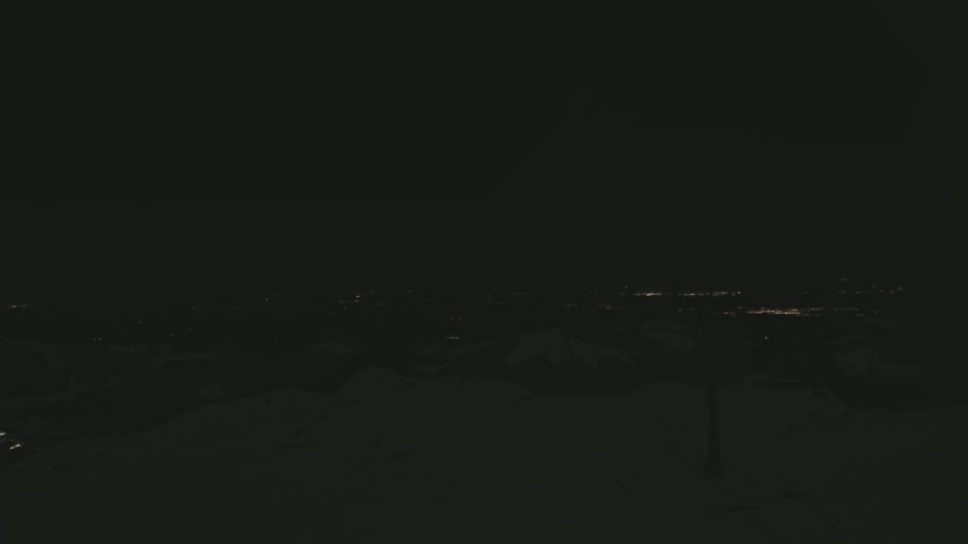 A nighttime view of a snow-covered landscape with a distant city's lights visible in the darkness.
