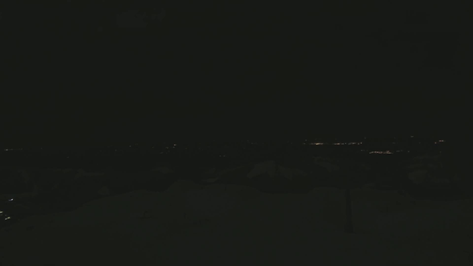 A dark, snowy landscape at night with a distant city's lights visible on the horizon.