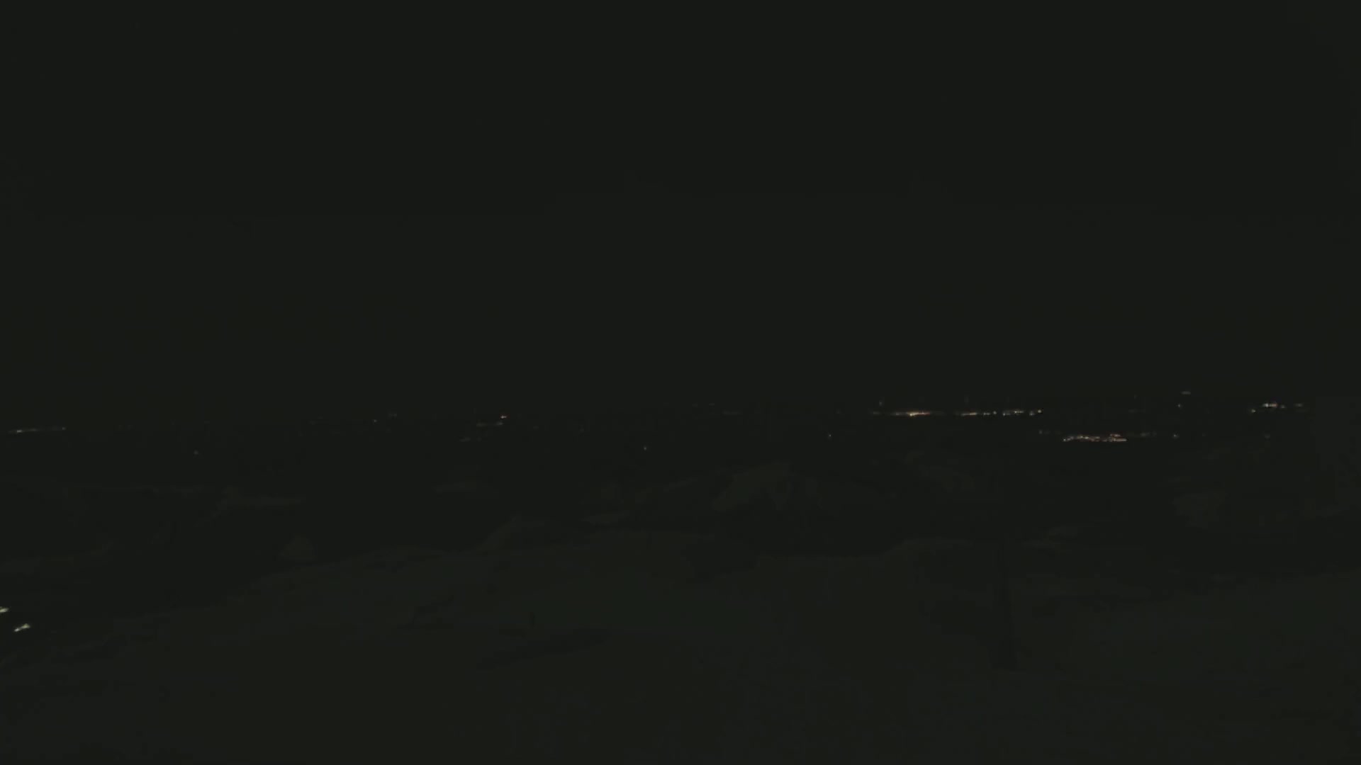 A dark, nighttime landscape shows a distant line of faint city lights in the background.