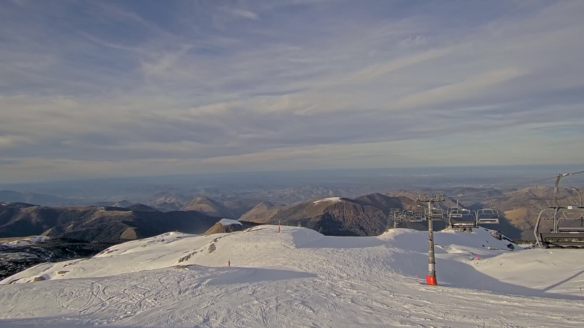 A snowy mountaintop ski resort with a chairlift overlooks a vast valley under a partly cloudy sky.