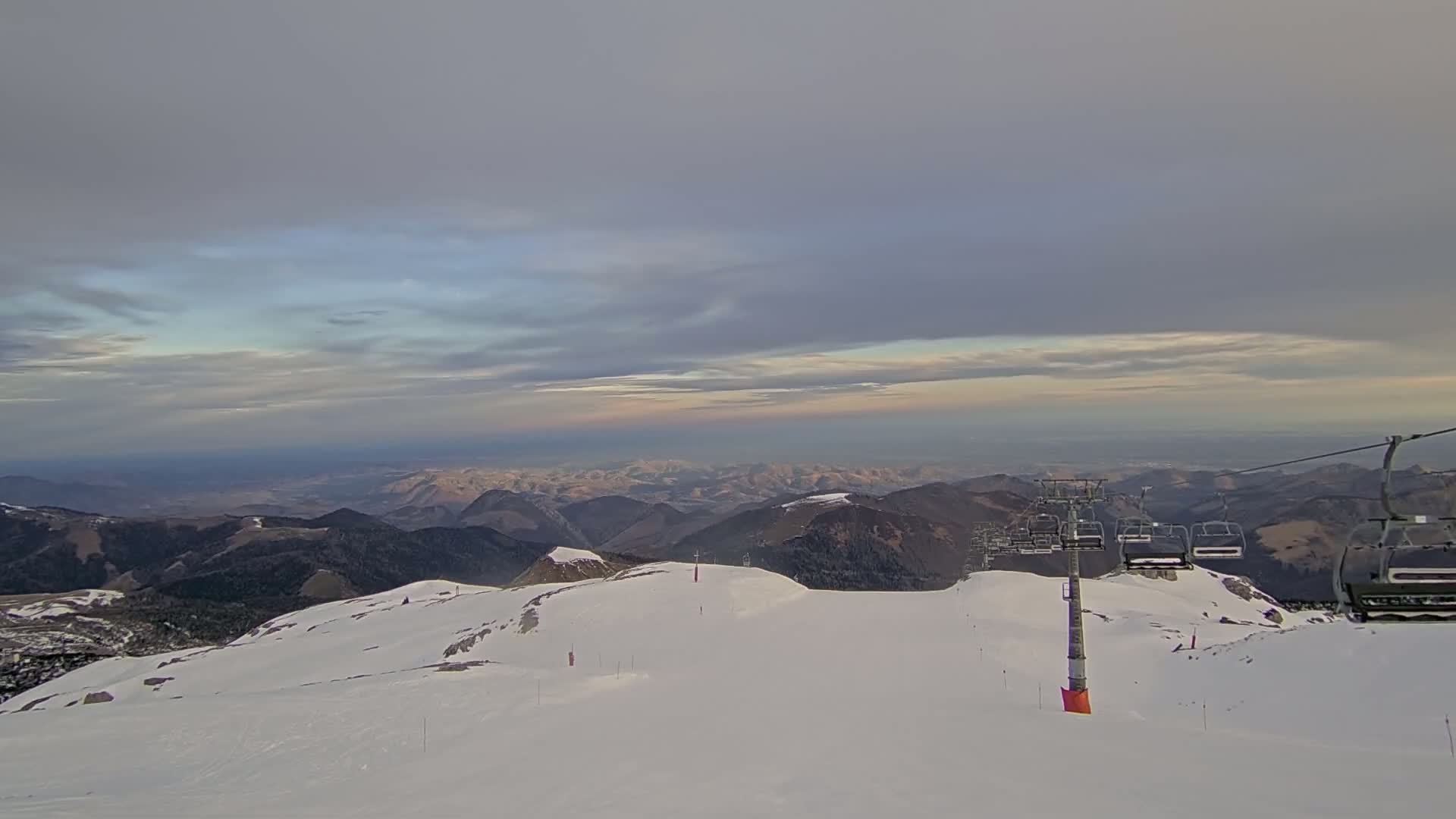 A snow-covered mountaintop with a chairlift and a view of distant mountains under a cloudy sky.