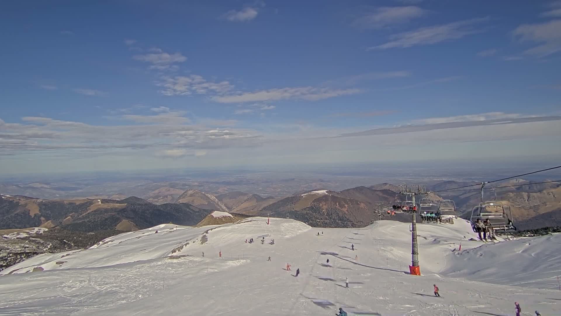 A snow-covered mountain slope with a chairlift and skiers, overlooking a vast, hazy valley under a mostly clear, bright blue sky.