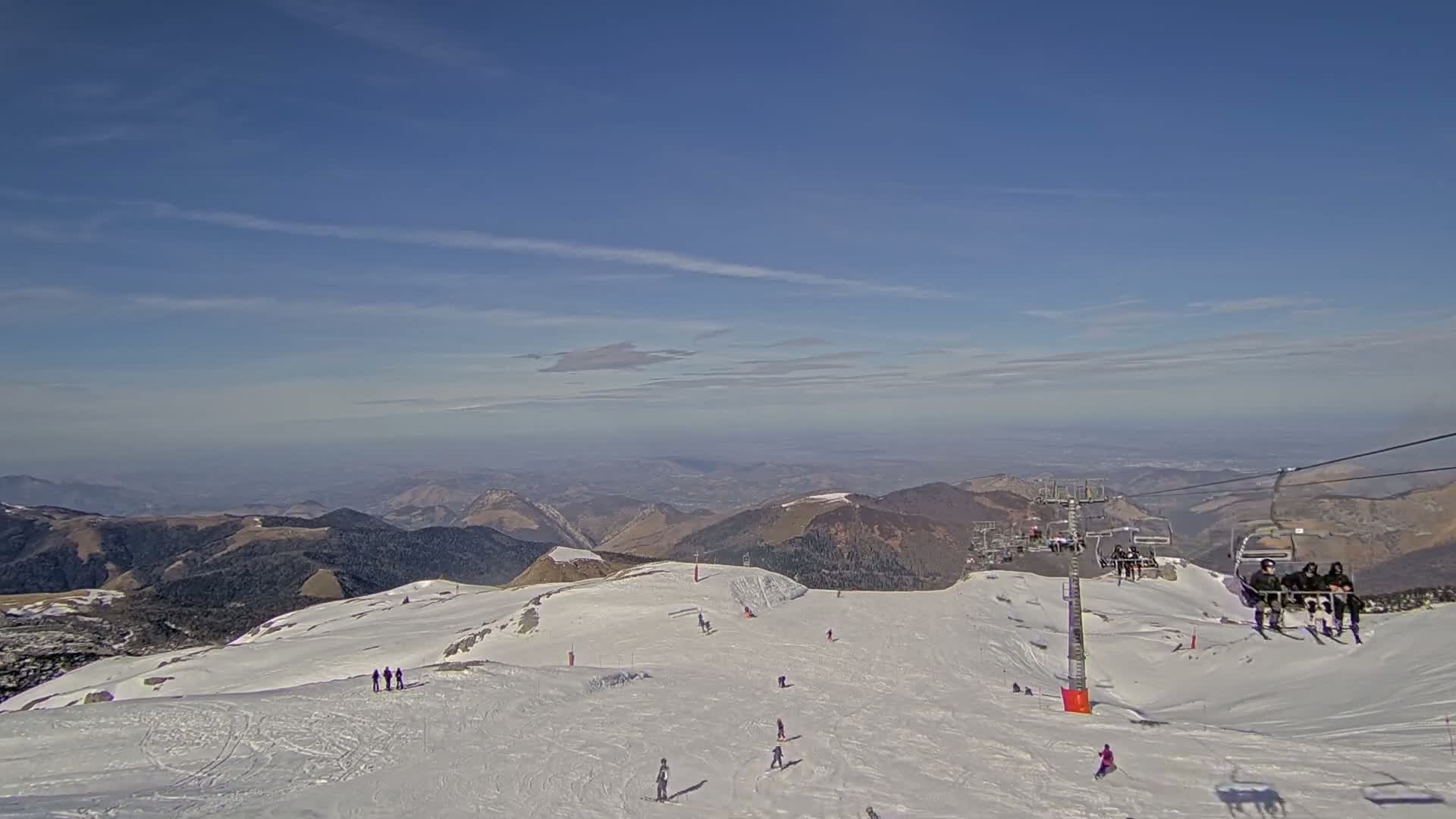 A snowy mountaintop ski slope with several skiers and snowboarders, chairlifts, and a clear, sunny sky overlooking distant mountain ranges.