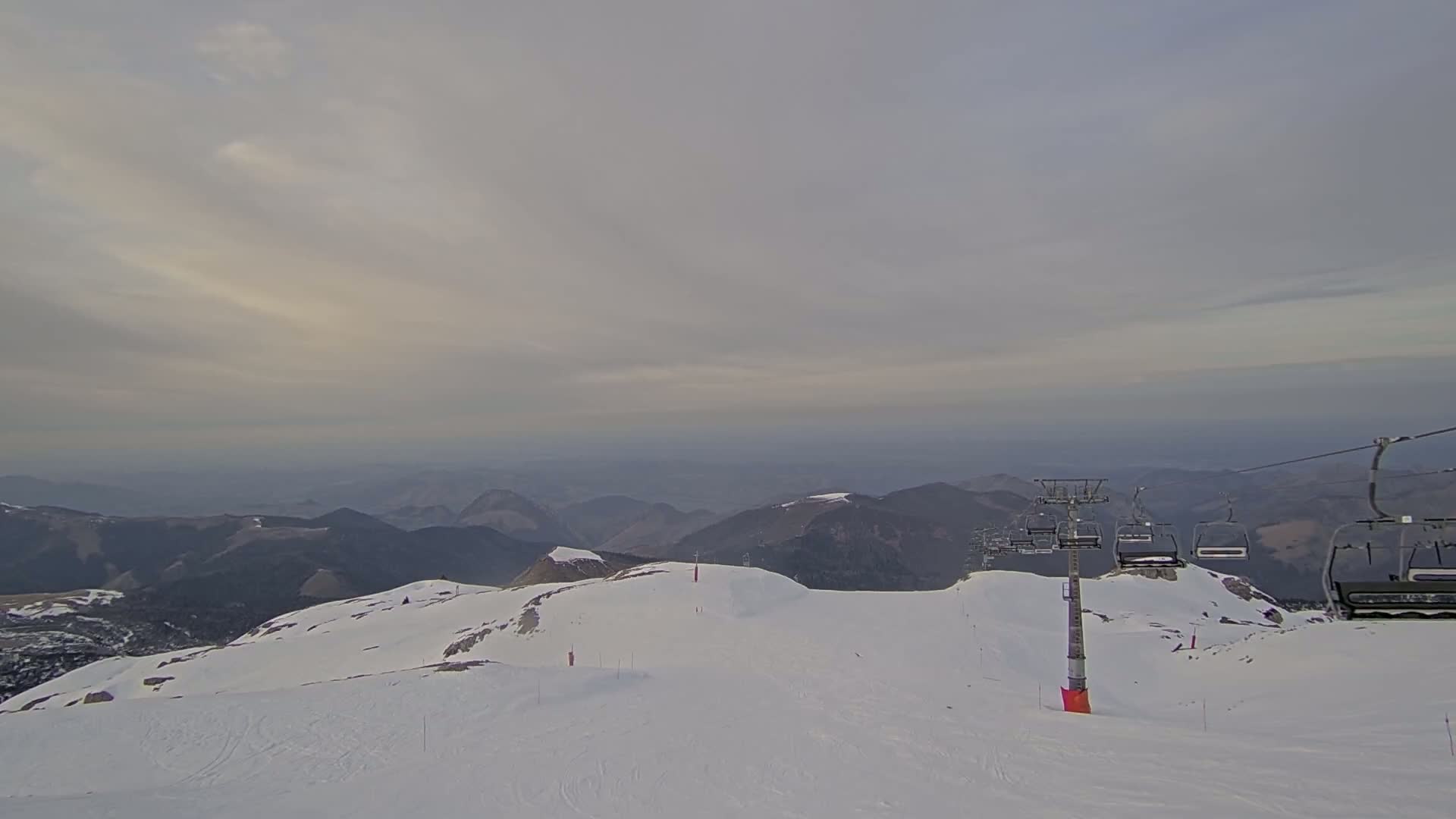A snow-covered mountaintop with a chairlift and distant mountain ranges under a cloudy sky.