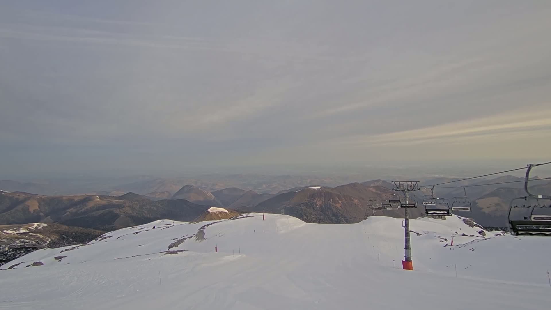 A snow-covered mountain slope with a chairlift and distant, hazy mountains under a mostly cloudy sky.
