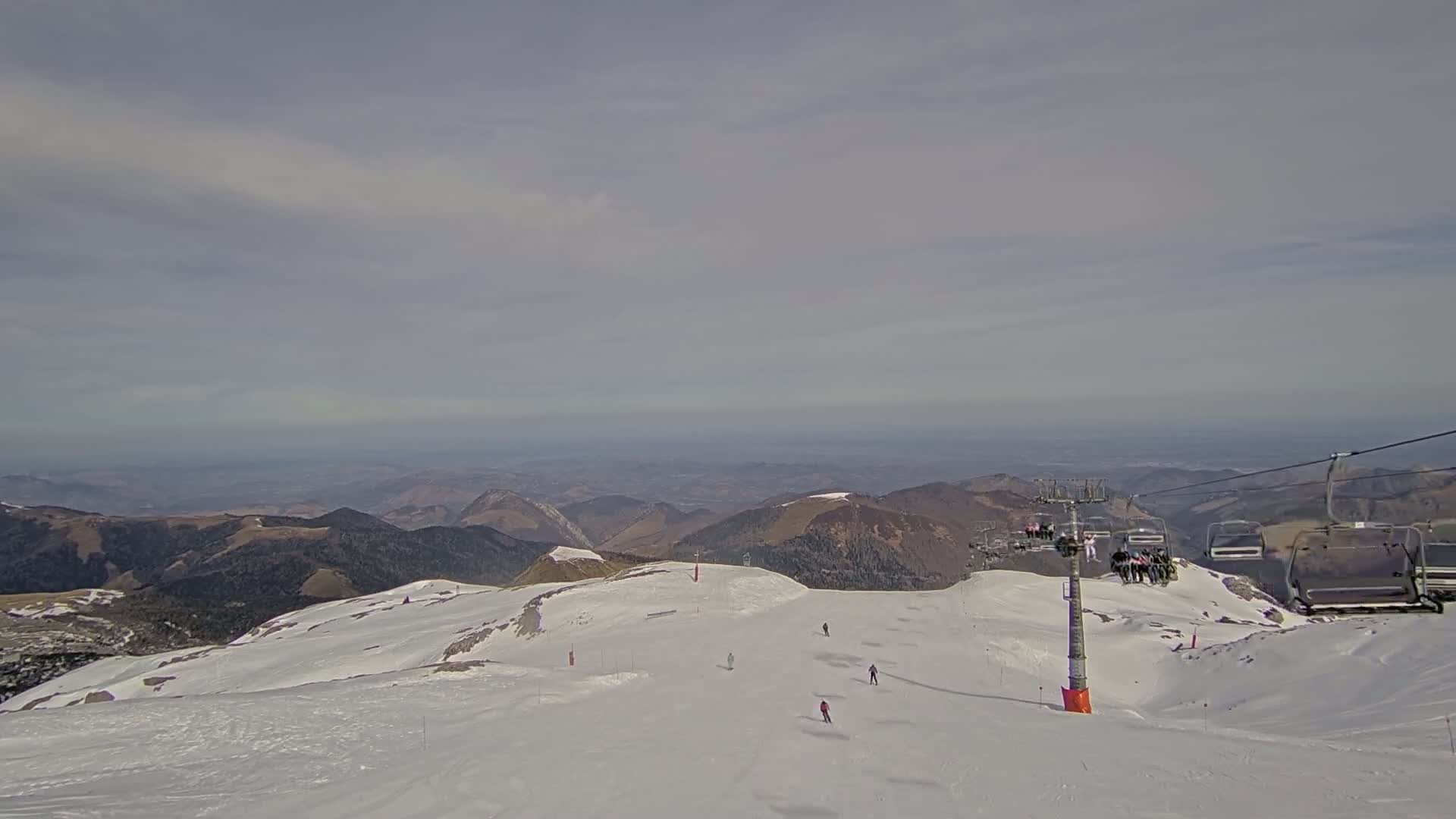 A ski slope with a chairlift and several skiers is visible against a backdrop of distant mountains under a cloudy sky.