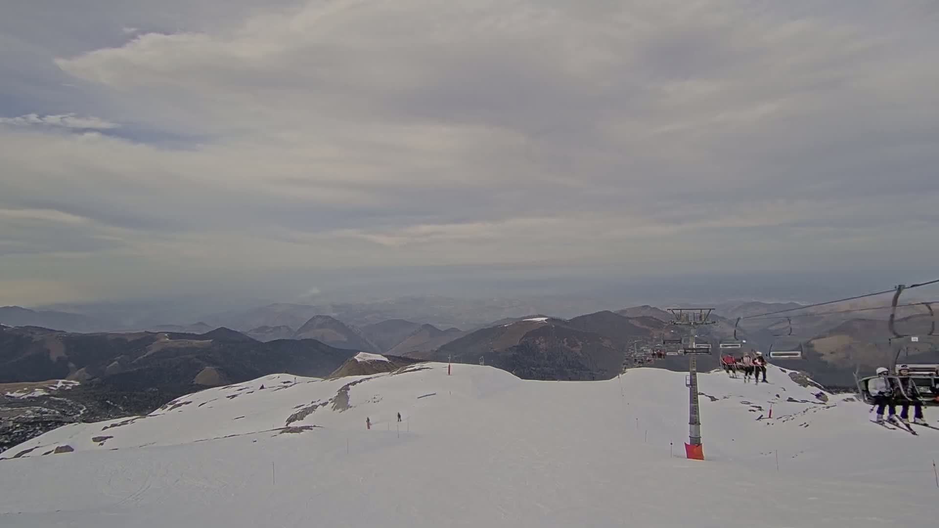A snow-covered mountaintop with a chairlift and a few skiers, under a cloudy sky.