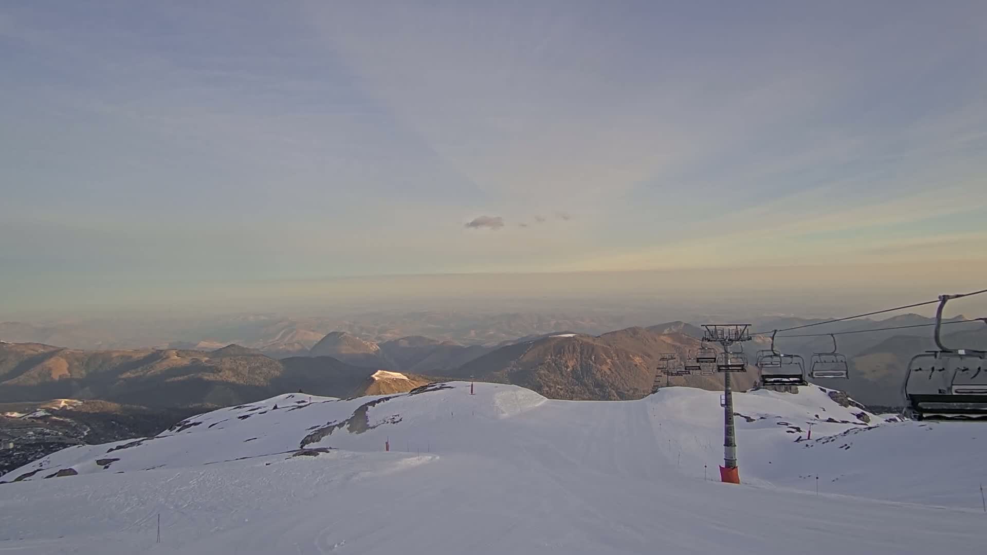 A snow-covered mountain slope with a chairlift extending towards a hazy, mountainous horizon under a mostly clear sky.