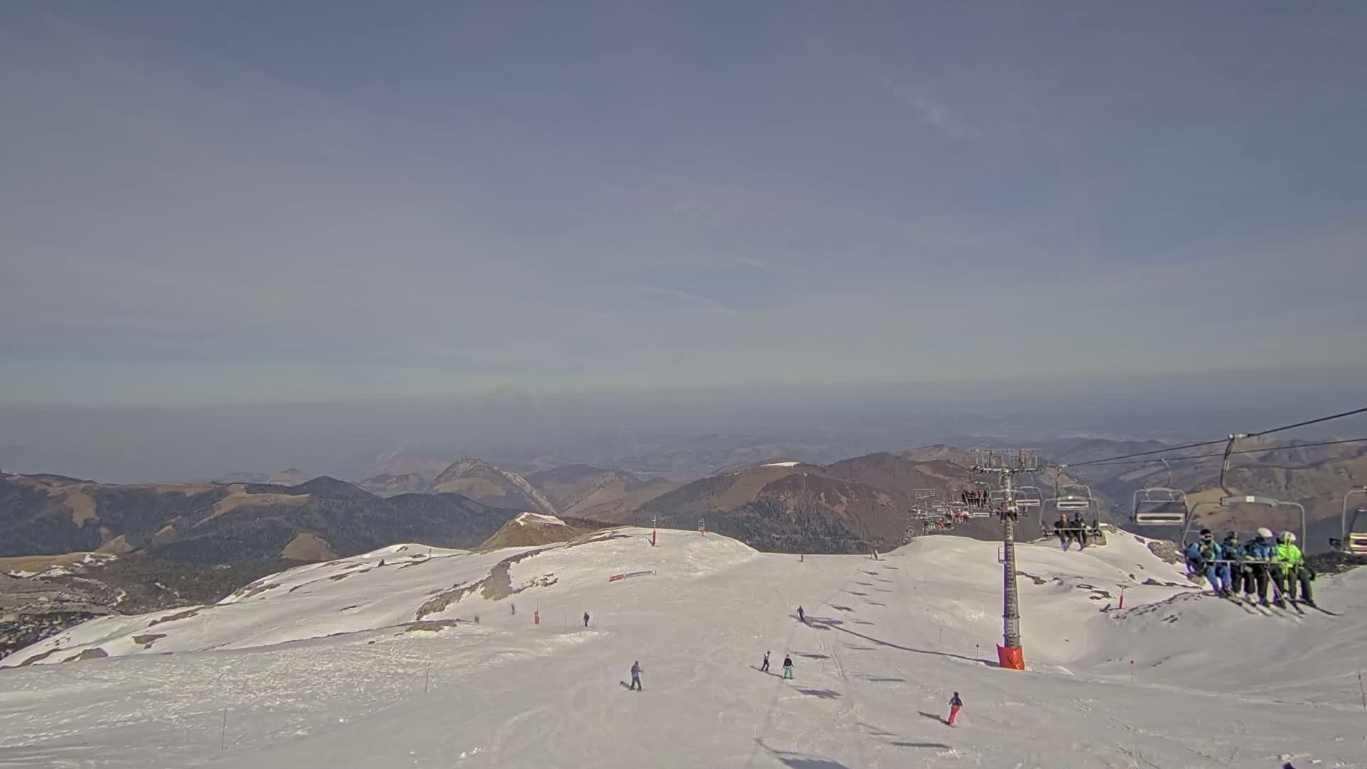 A mostly clear, hazy day reveals a snow-covered mountain slope with skiers and a chairlift against a backdrop of distant mountains.