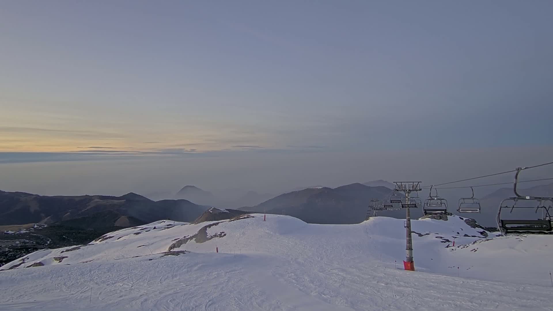 A chairlift stands on a snow-covered mountaintop at sunset, overlooking a valley shrouded in fog.
