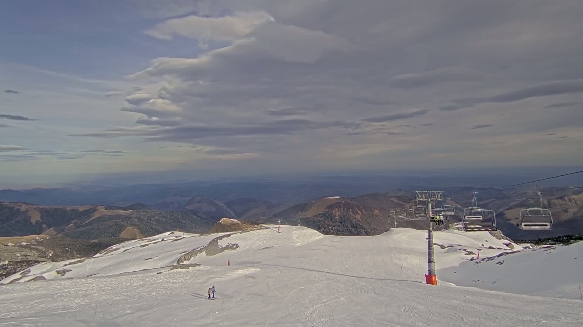 A mostly cloudy sky overlooks a snowy mountaintop ski slope with a chairlift and two skiers.