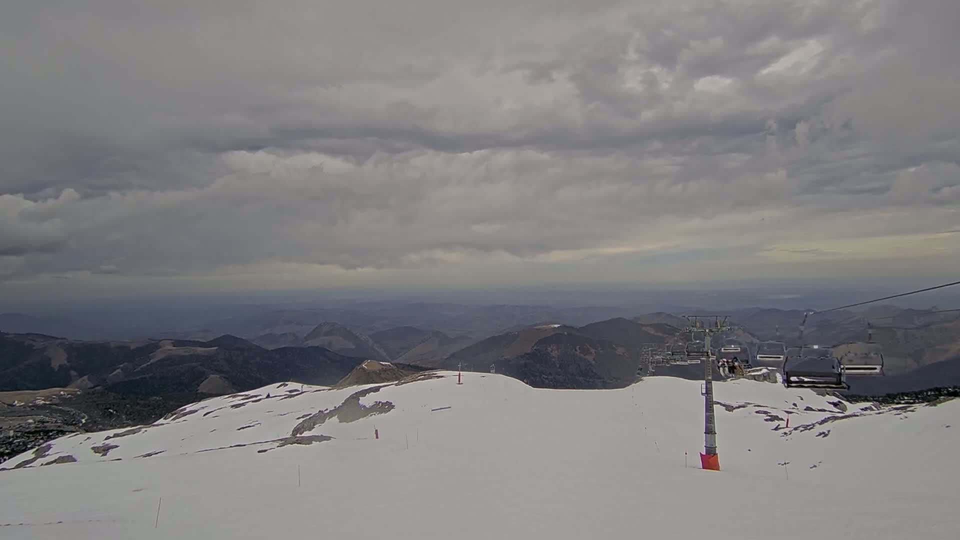 A mostly cloudy day reveals a snow-covered mountaintop with a chairlift and distant mountain ranges.