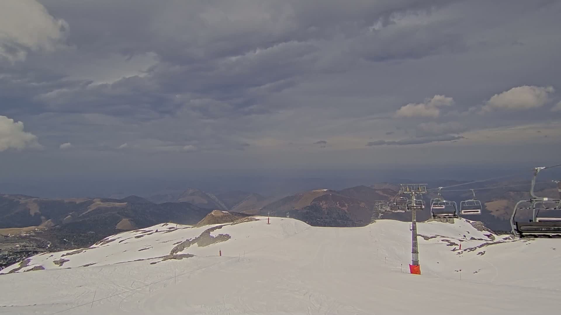 A snowy mountaintop with a chairlift and overcast skies overlooks a distant mountain range.