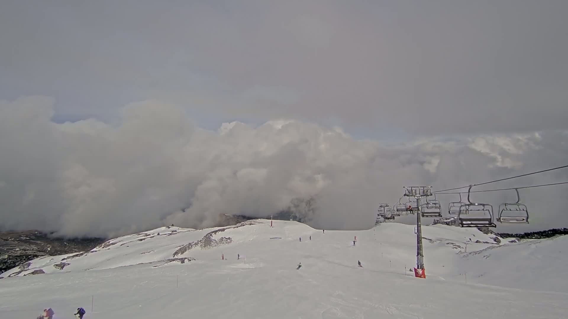 A snowy mountain slope with a chairlift and several skiers under a cloudy sky.