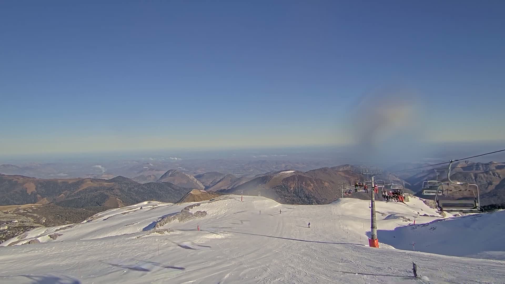 A snow-covered mountain slope with a chairlift and several skiers under a clear blue sky.