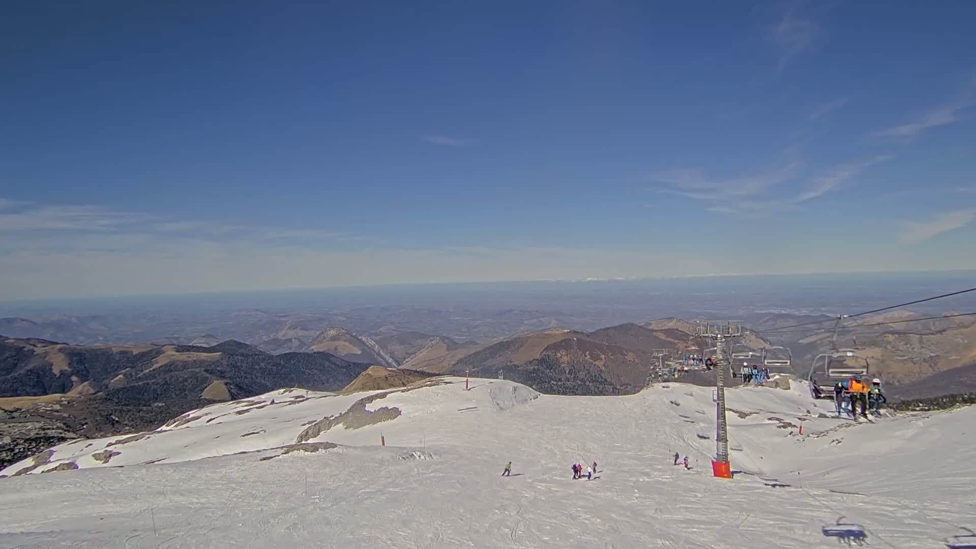 A sunny, snow-covered mountaintop ski slope with a chairlift and several skiers visible, overlooking a vast expanse of distant mountains and valleys under a clear blue sky.