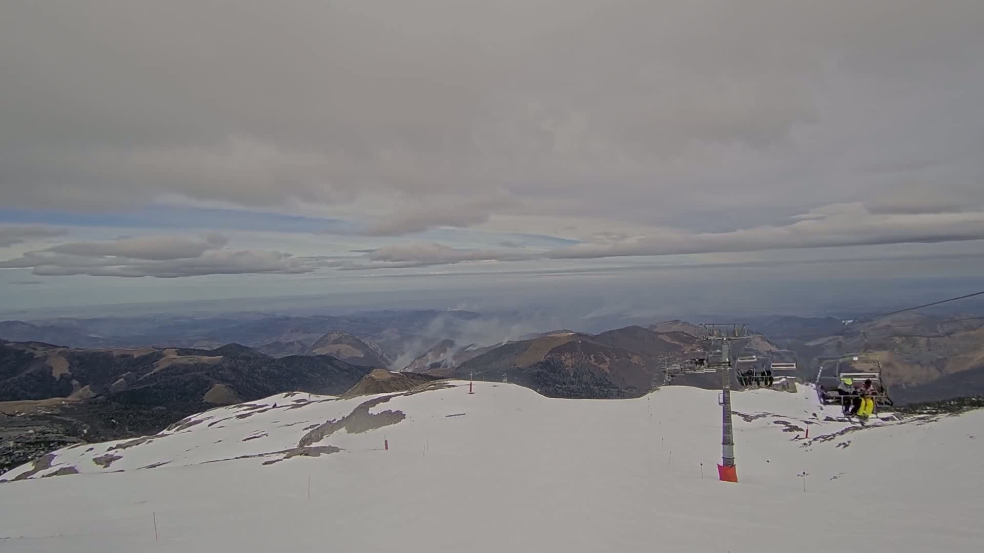 A snow-covered mountain peak with a chairlift and distant hazy mountains under a cloudy sky.