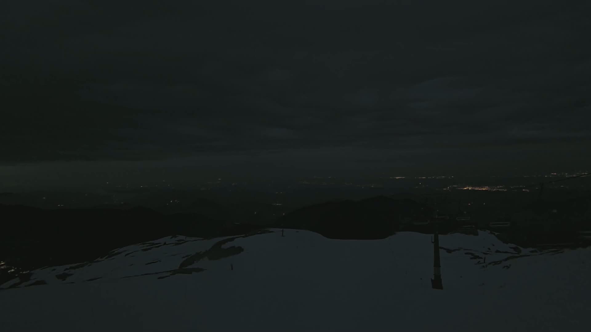 A snow-covered mountain slope at night overlooks a distant city illuminated by faint lights under a dark, cloudy sky.