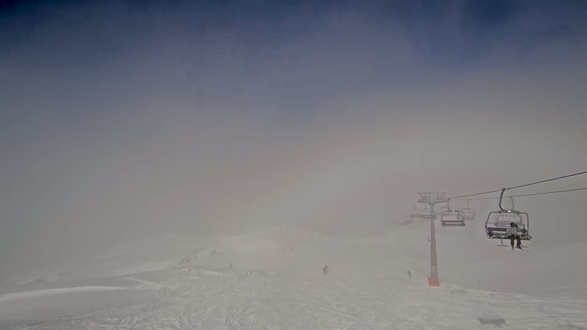 A ski lift with several occupied chairs ascends a snow-covered slope shrouded in thick fog.
