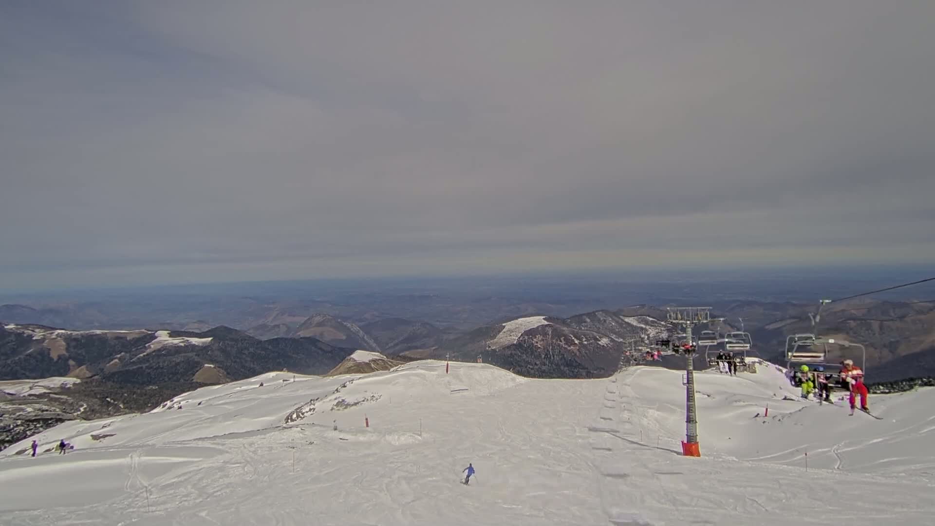 A snowy mountaintop ski slope with a chairlift and several skiers, overlooking a vast, hazy valley under a cloudy sky.