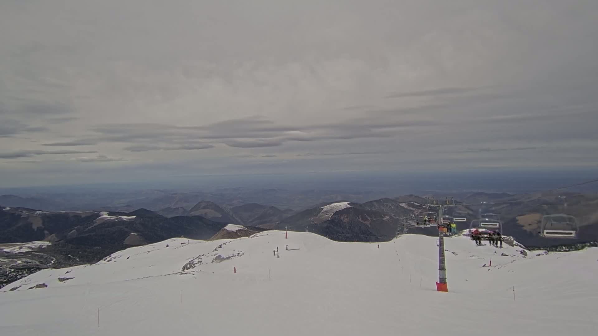 A snow-covered mountaintop with a chairlift and several skiers visible under an overcast sky.