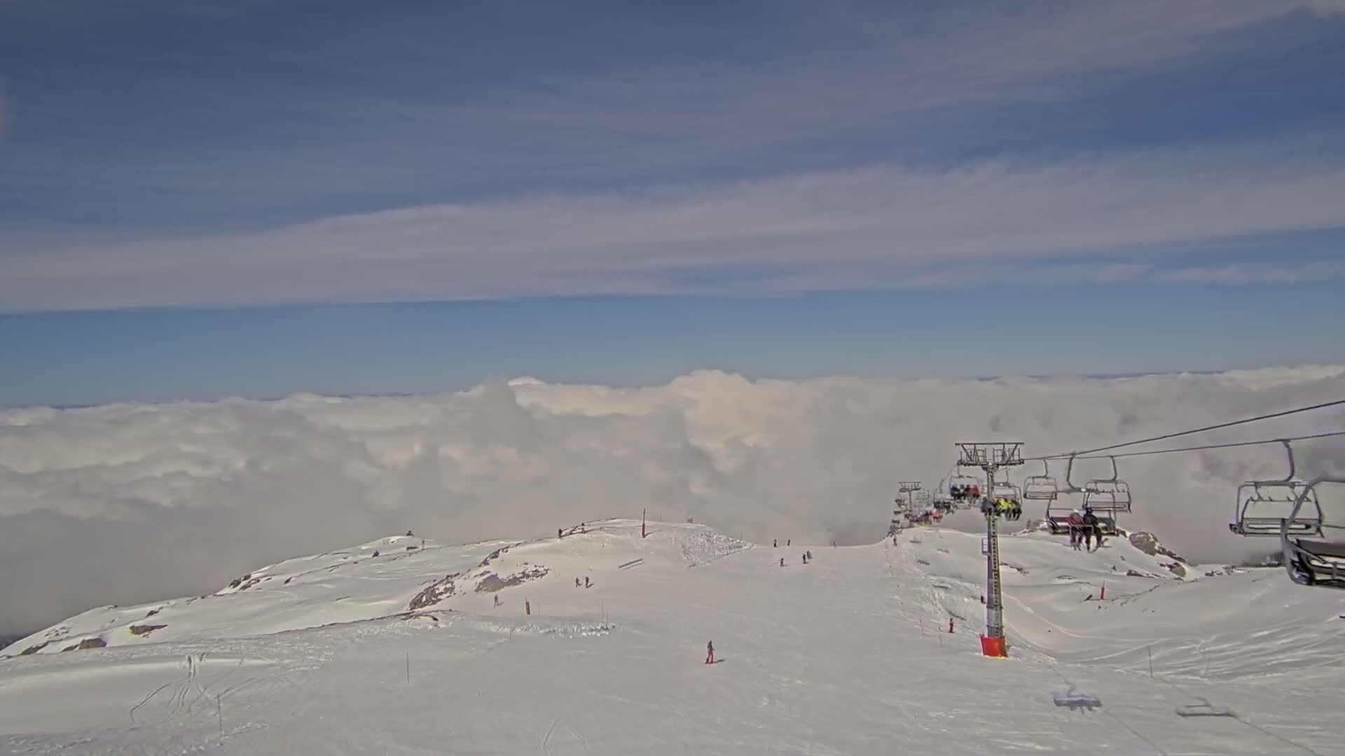 A snowy mountaintop ski slope with a chairlift carrying skiers overlooks a sea of clouds under a mostly clear blue sky.