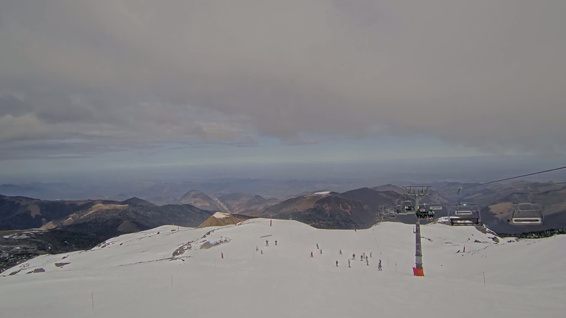 A ski slope with several skiers and a chairlift is visible, overlooking a hazy mountainous landscape under a cloudy sky.