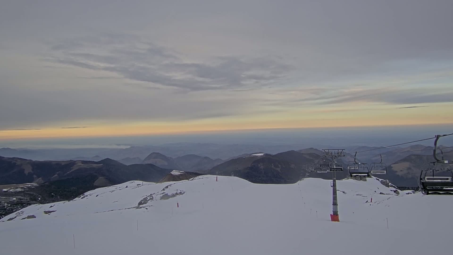 A snow-covered mountaintop at sunrise overlooks a valley shrouded in fog, with a chairlift visible in the foreground.