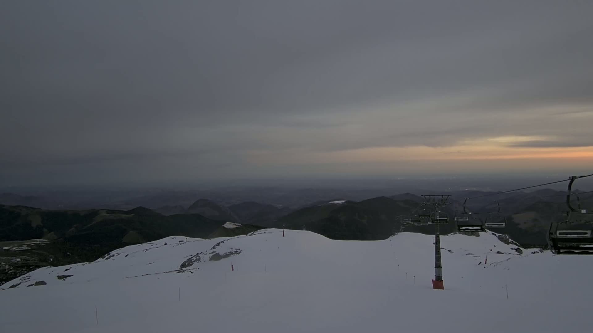 A snow-covered mountaintop with a chairlift, under a mostly cloudy sky with a hint of sunset in the distance.