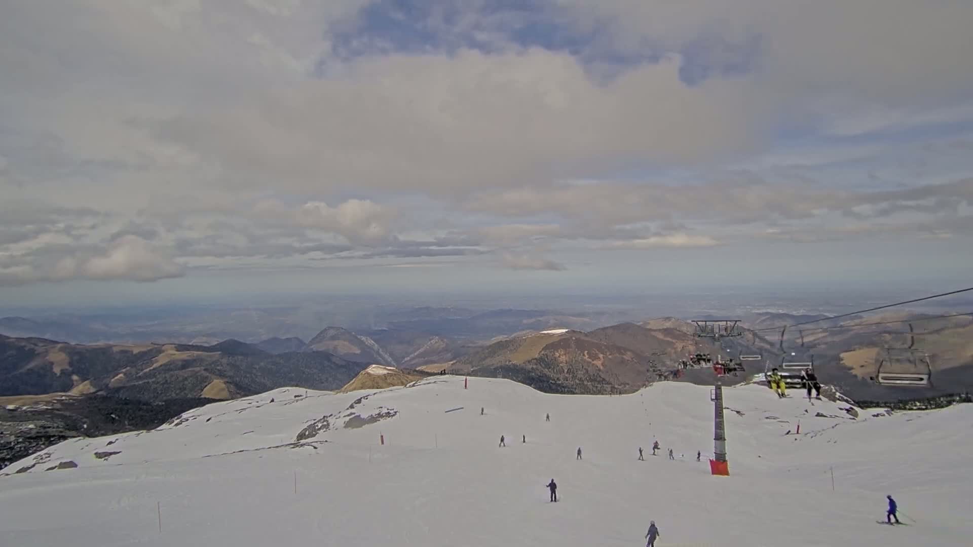 A mostly cloudy sky overlooks a snow-covered mountain slope with a chairlift and several skiers and snowboarders.