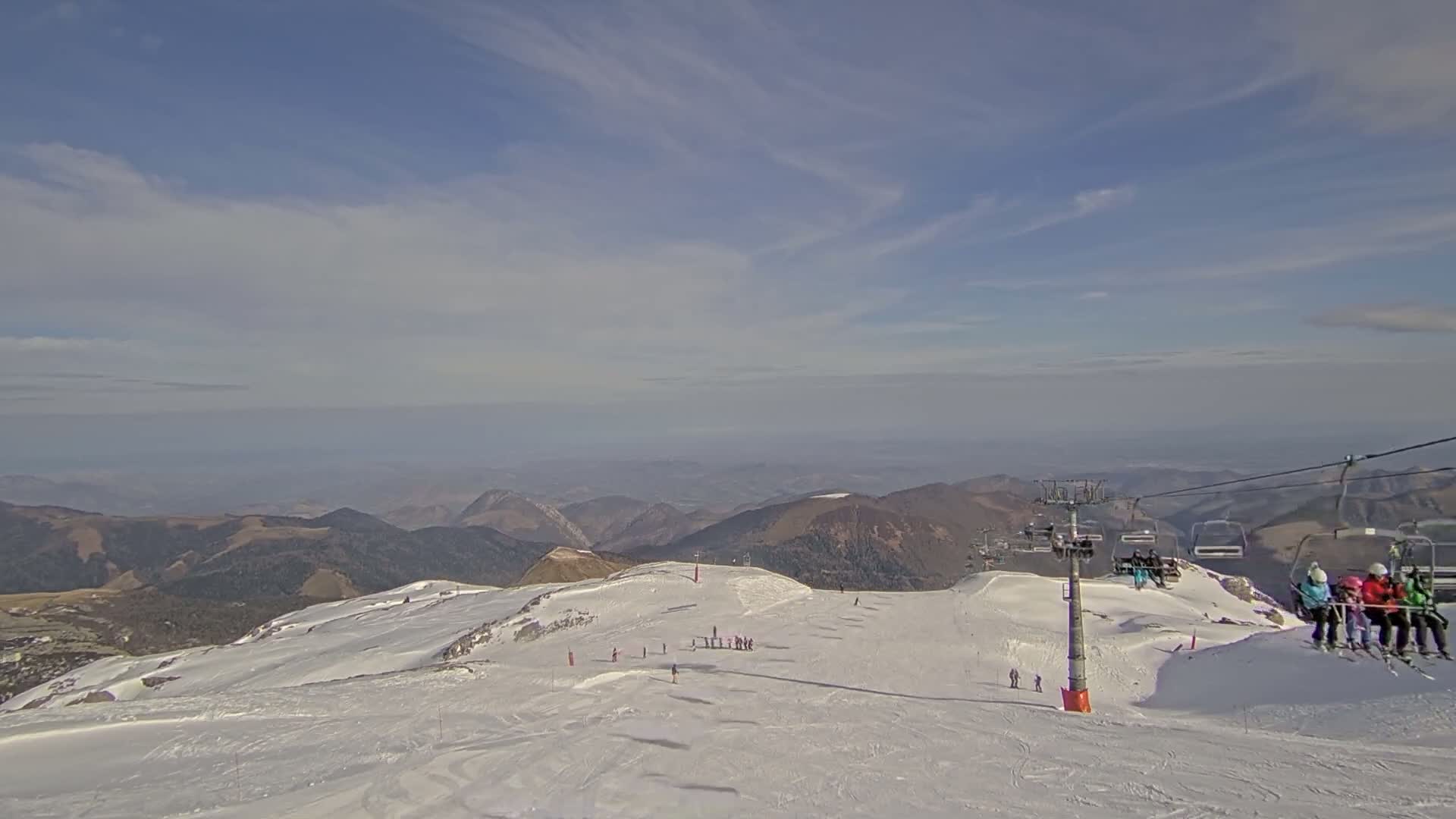A snow-covered mountaintop ski slope with a chairlift and scattered skiers under a mostly clear, bright sky overlooking a hazy valley.