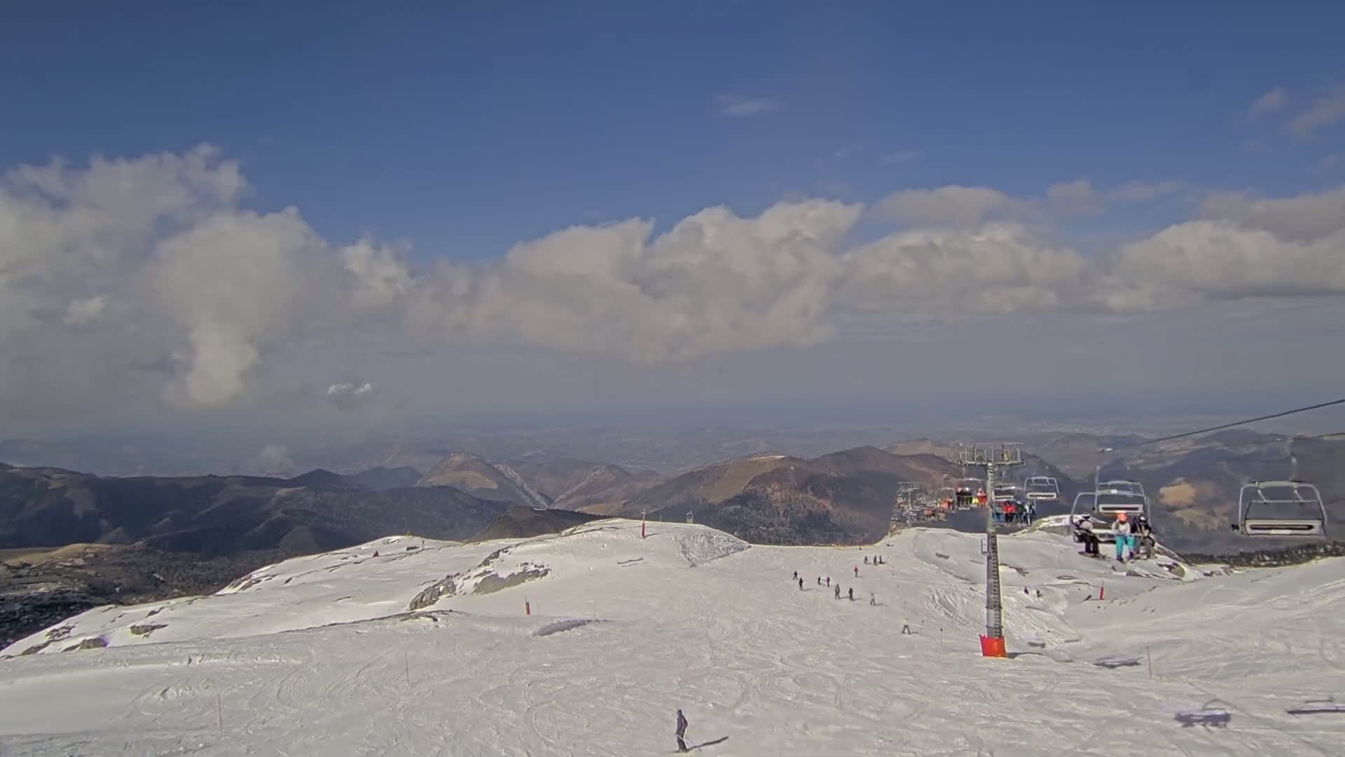 A snowy mountain slope with a chairlift and several skiers under a partly cloudy sky.