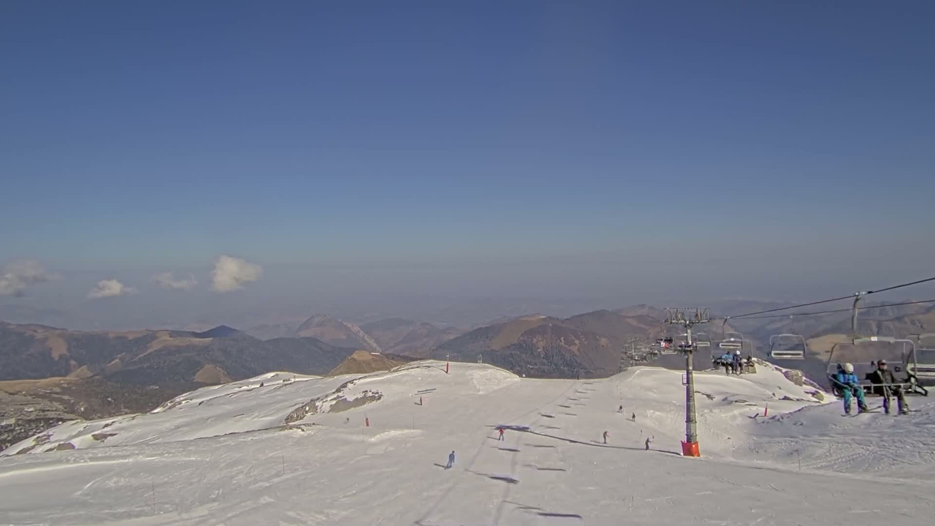 A ski slope with a chairlift and several skiers is visible against a backdrop of distant mountains under a clear blue sky with a few wispy clouds.