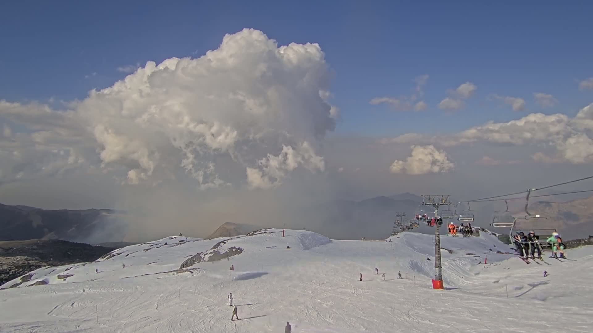 A snowy mountain slope with skiers and a chairlift is visible under a partly cloudy sky, with a large cumulus cloud dominating the scene and hazy smoke or dust visible in the distance.