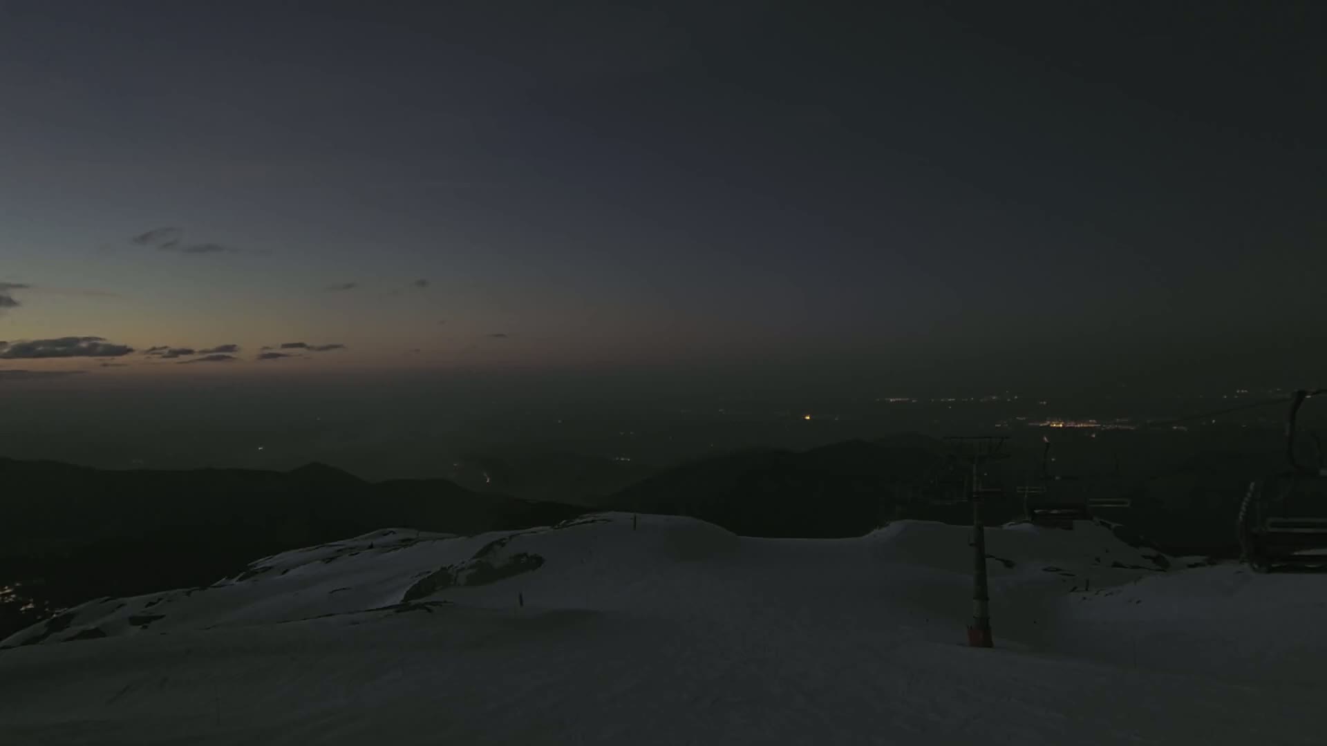 A snow-covered mountaintop at twilight, with a chairlift visible and distant city lights twinkling below.