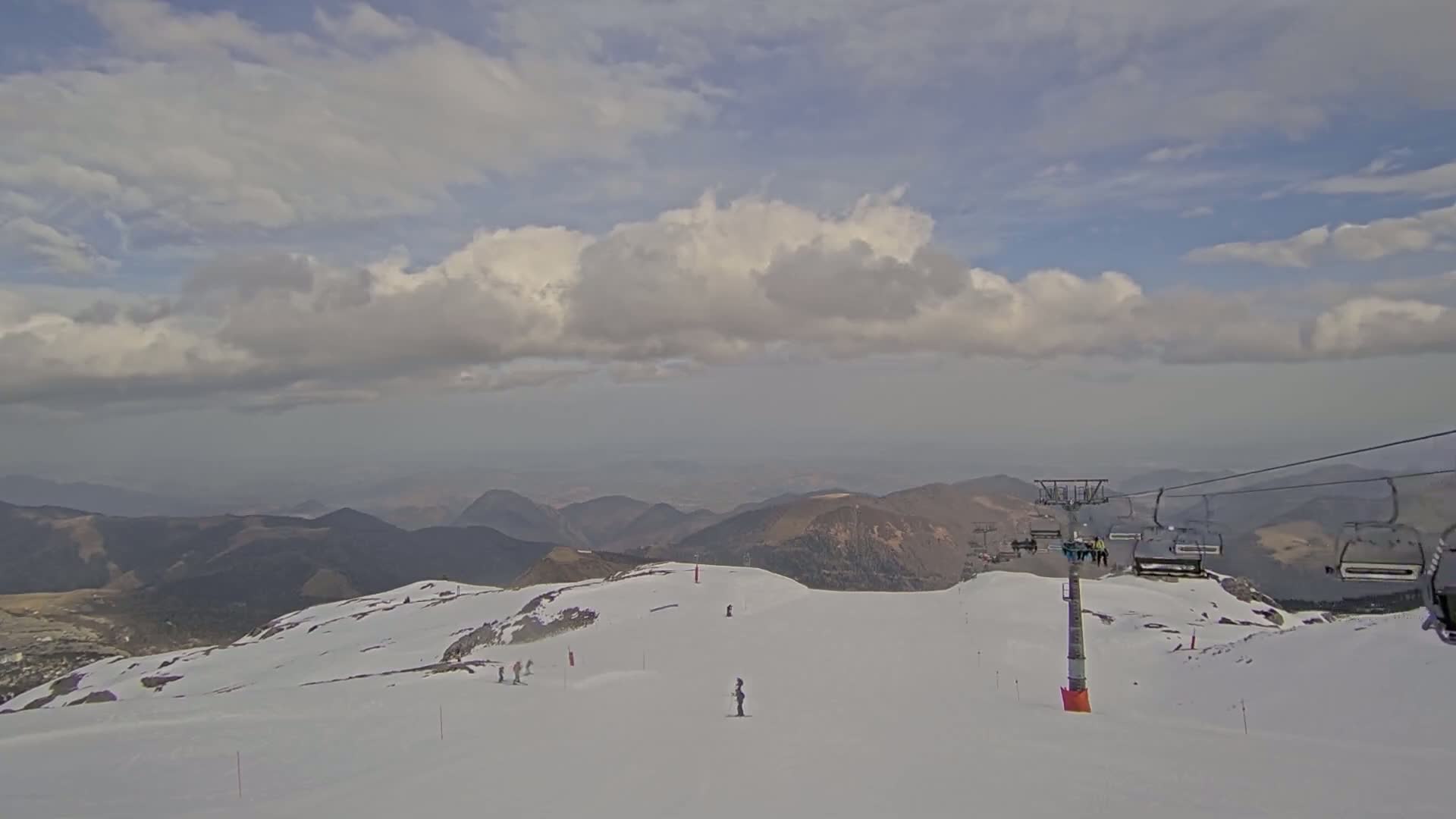 A mostly cloudy day reveals a snowy mountaintop ski slope with a chairlift and a few skiers, overlooking a range of distant mountains.