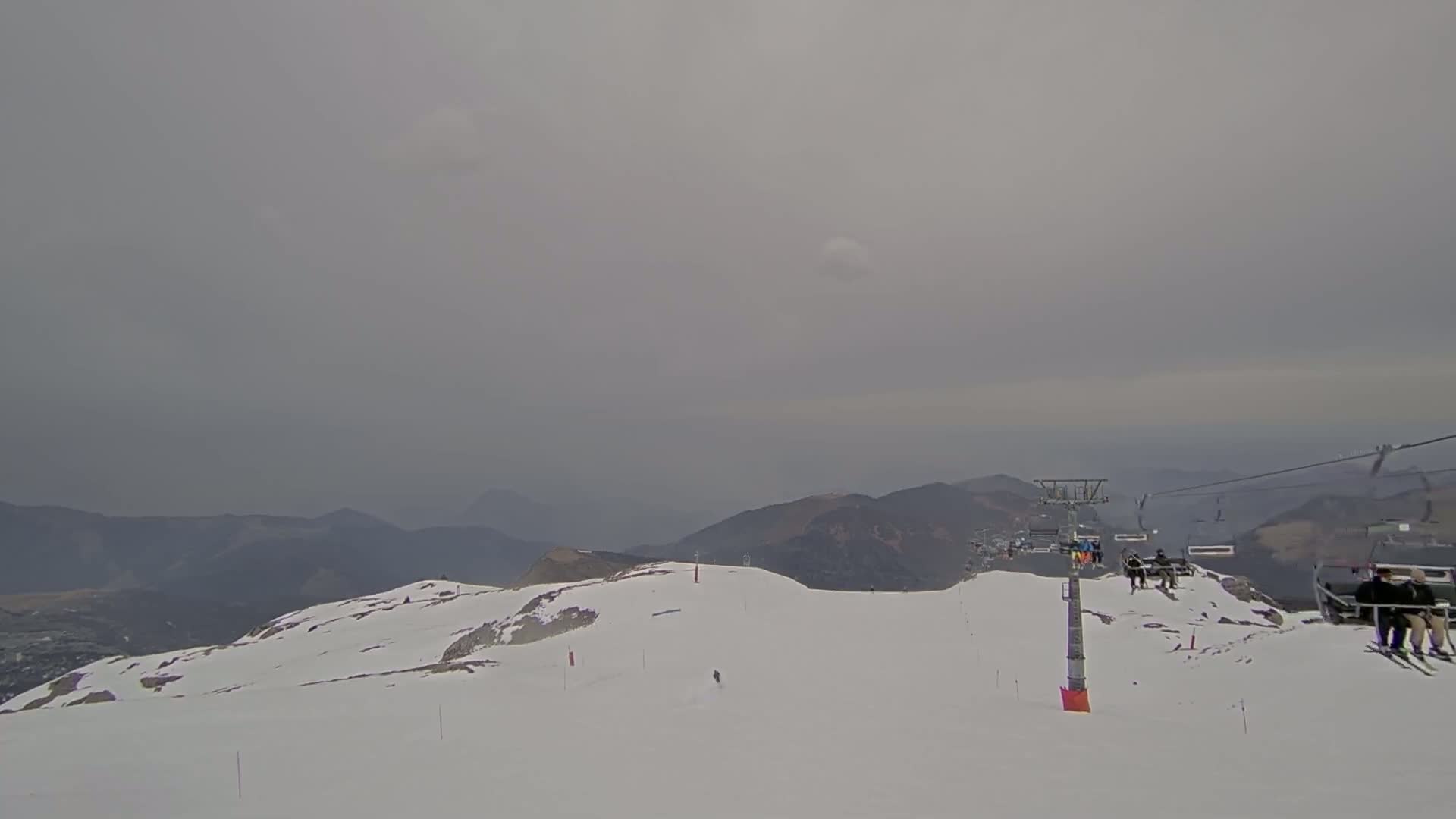 A snowy mountaintop scene under an overcast sky shows a ski lift with several people riding in the chairs, and a lone skier on a slope in the distance.