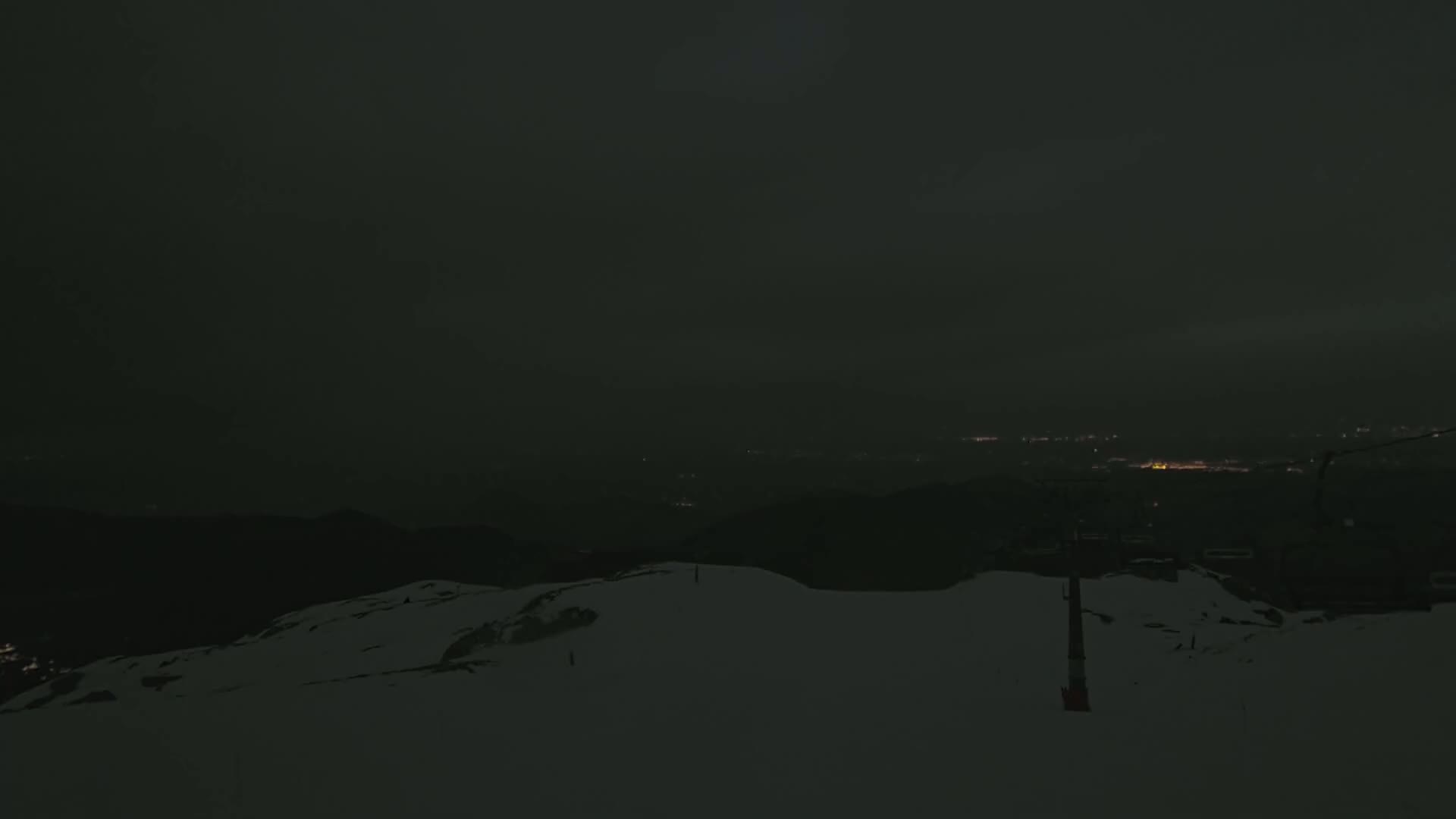 A snow-covered mountaintop at night overlooks a dimly lit valley under a dark, cloudy sky.