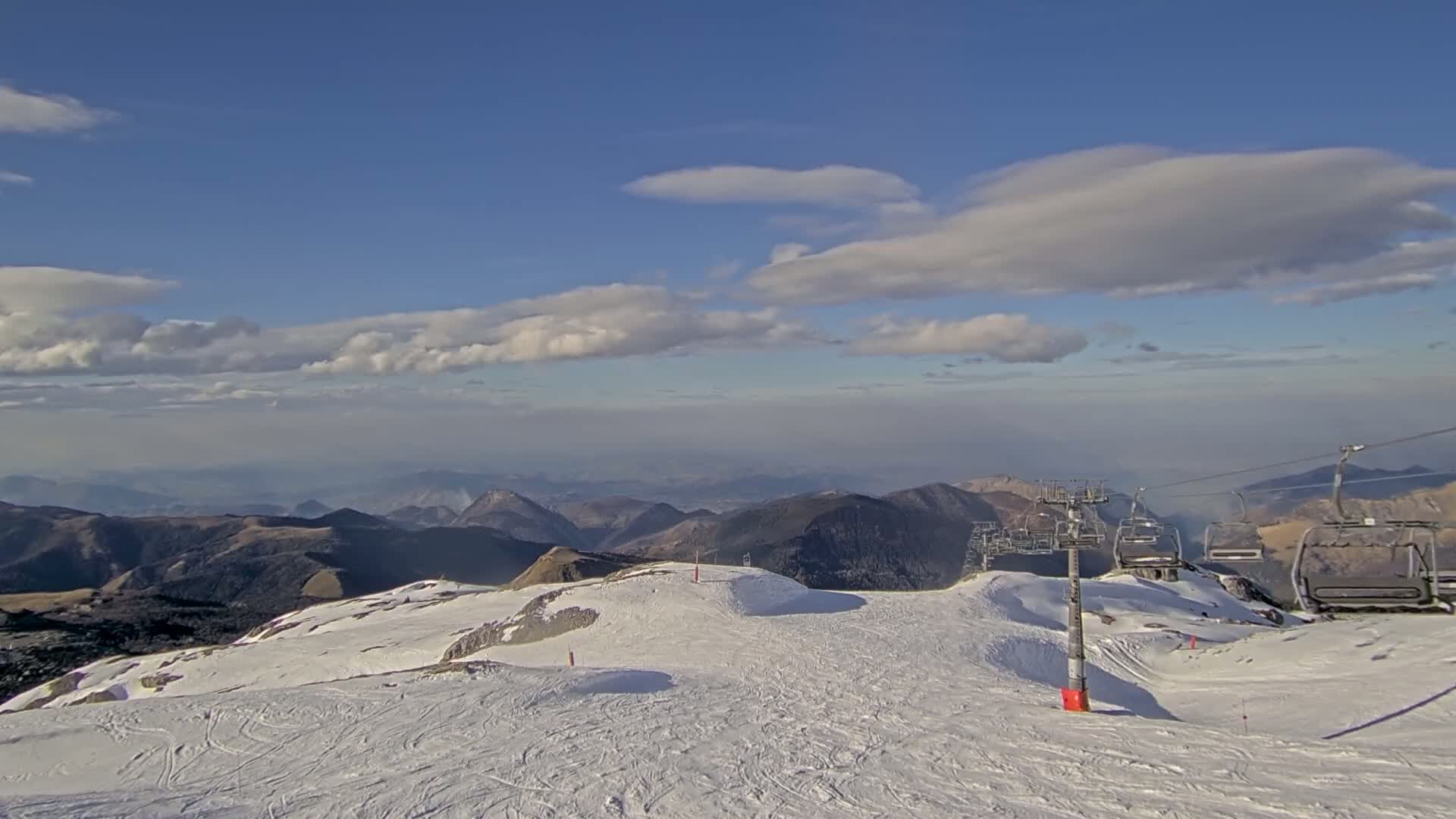 A snow-covered mountaintop with a chairlift and distant mountain ranges under a partly cloudy blue sky.
