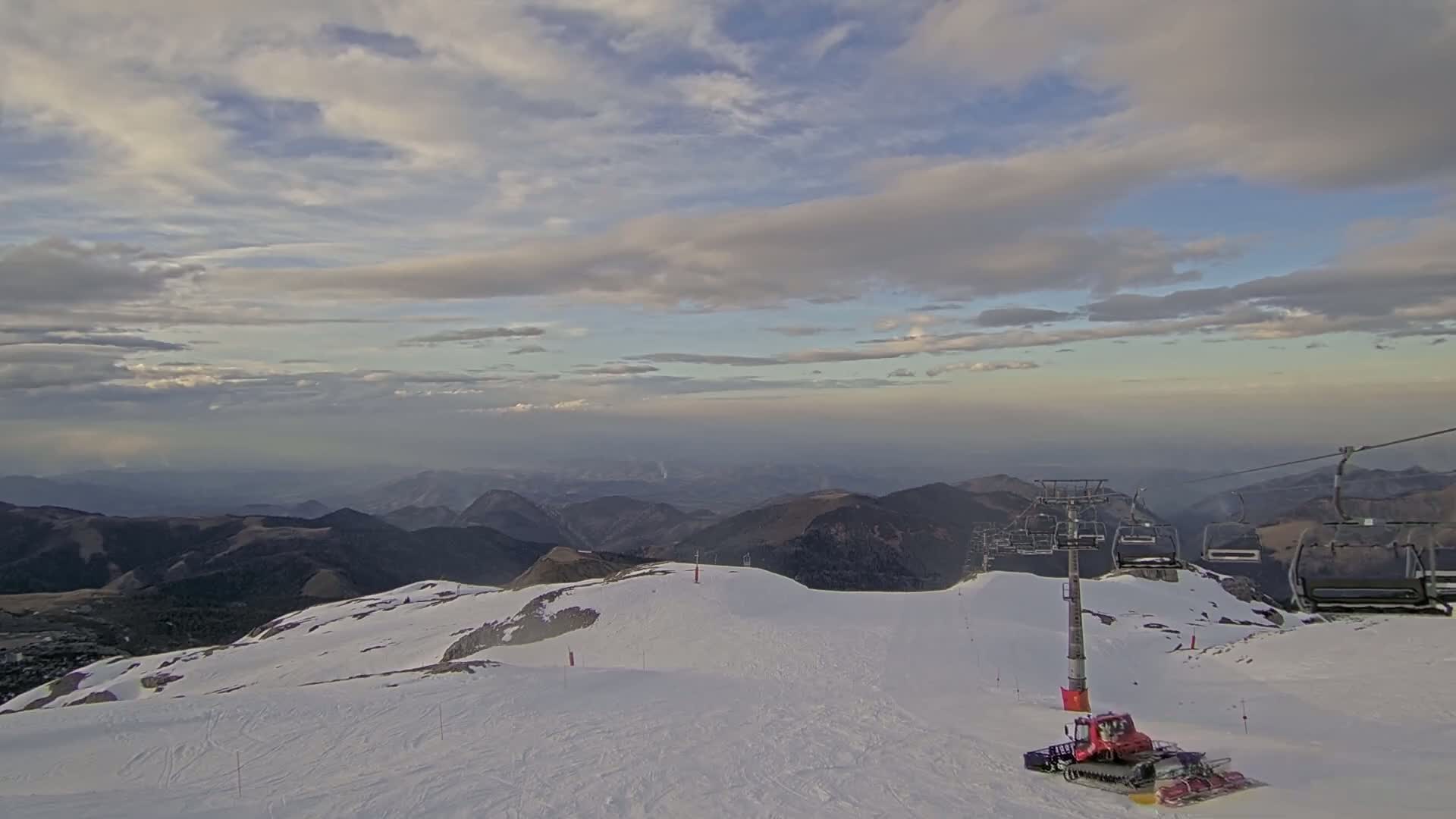 A snow-covered mountaintop with a ski lift and snow groomer under a partly cloudy sky.