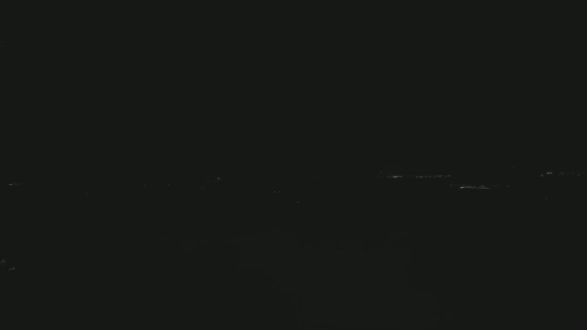 A nighttime long shot shows a dimly lit horizon with faint lights in the distance under a dark sky.