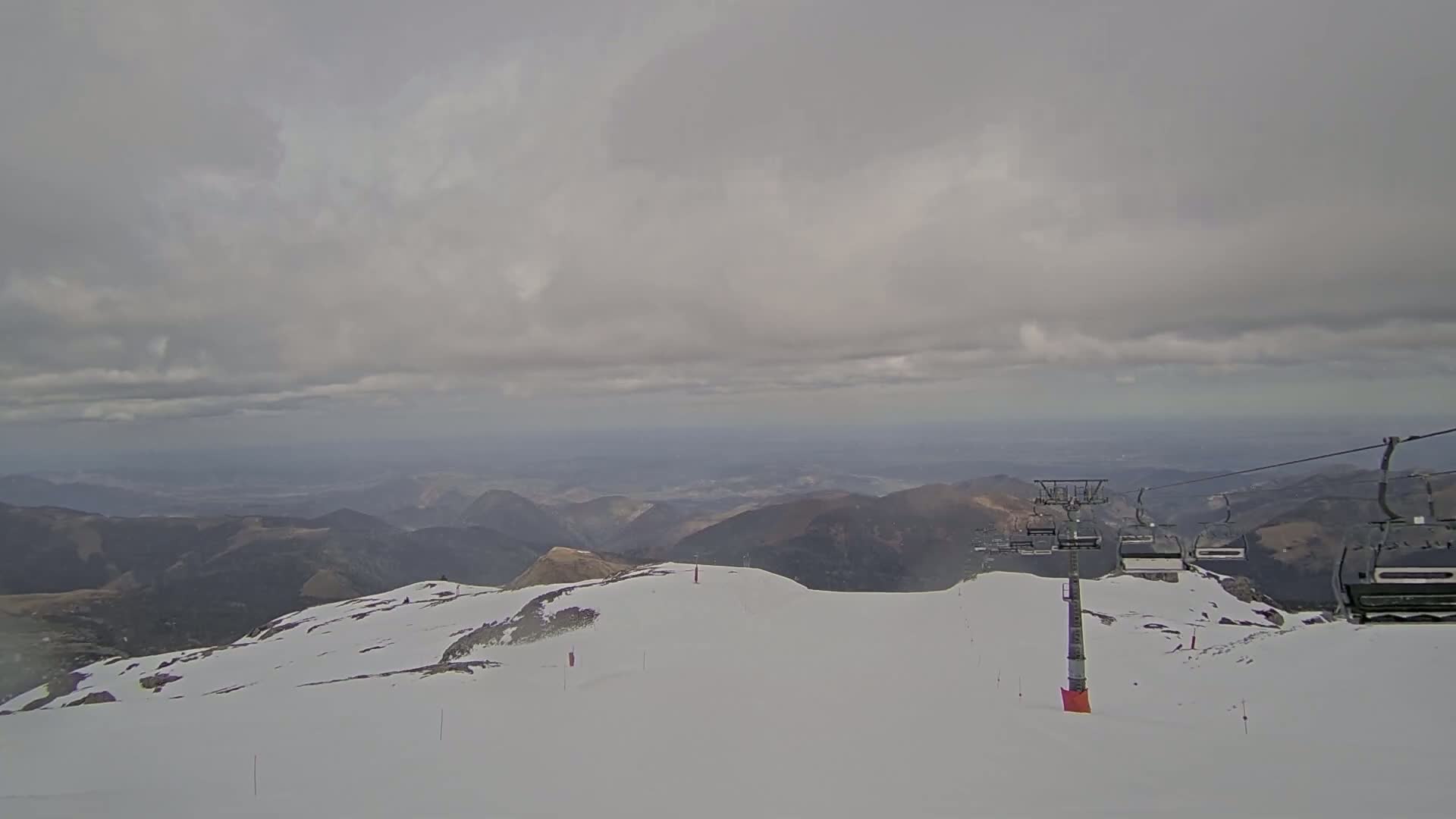 A snowy mountaintop with a chairlift and overcast skies overlooks a distant range of brown hills.