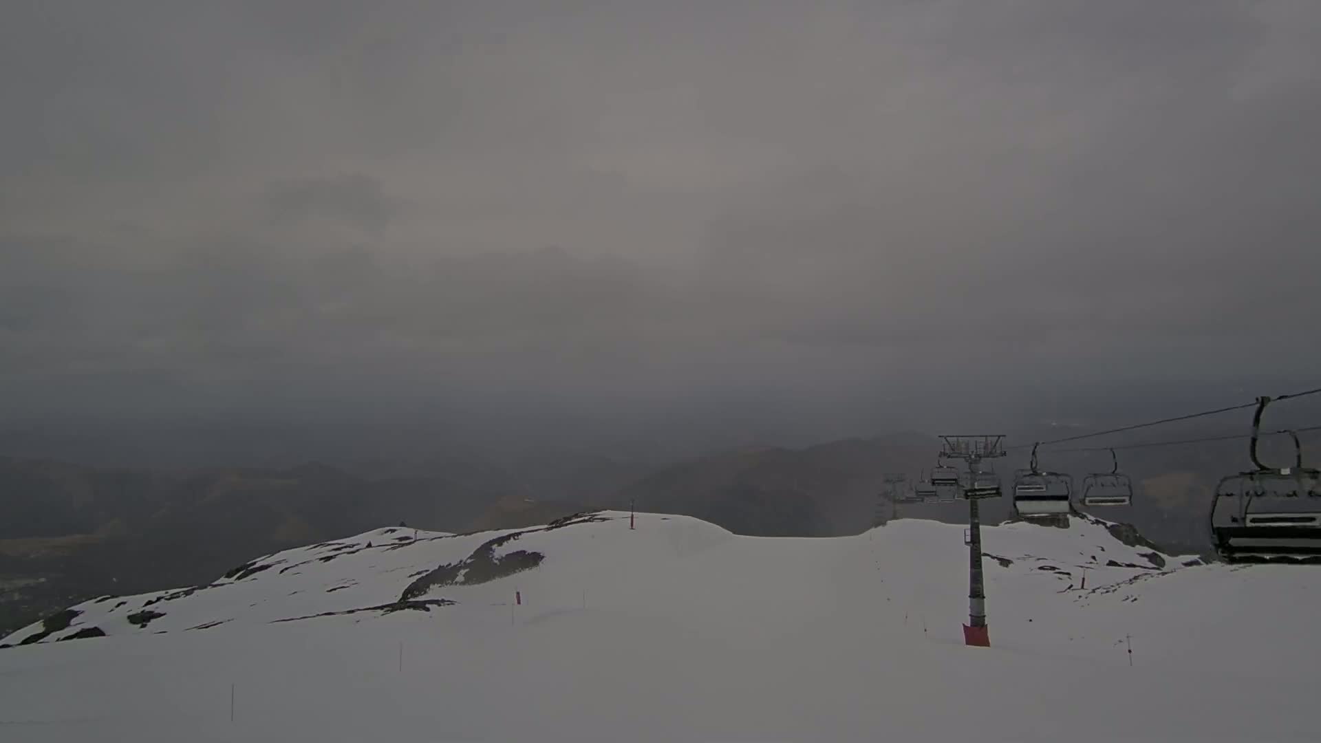 A snow-covered mountain landscape under a cloudy, overcast sky features a chairlift with several empty chairs.