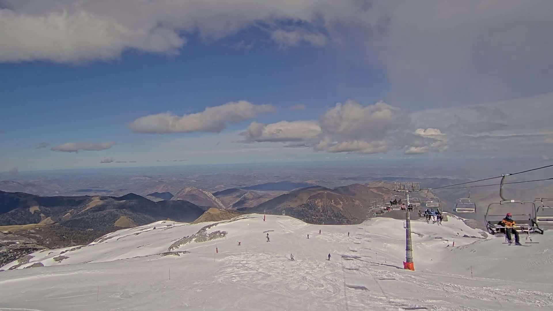 A snowy mountain slope with skiers and a chairlift, overlooking a vast valley under a partly cloudy sky.