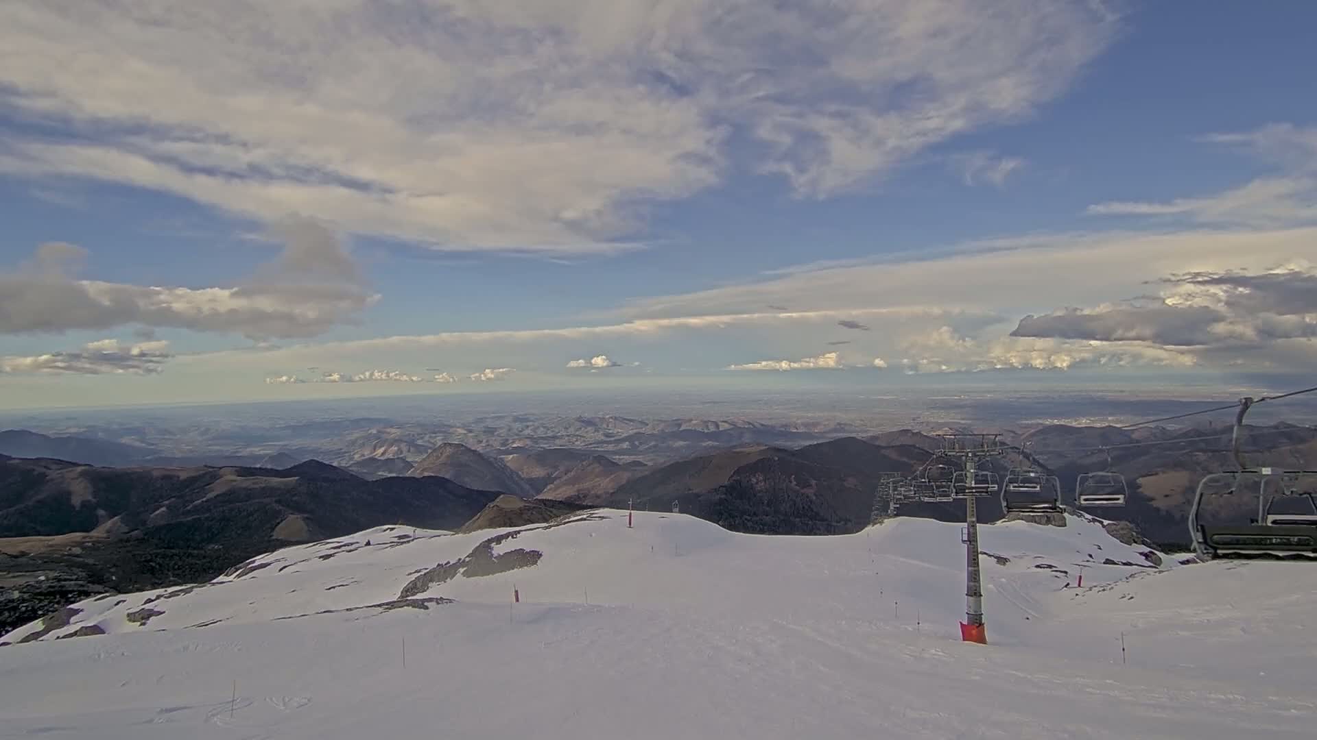 A snow-covered mountaintop with a chairlift overlooks a vast valley under a partly cloudy sky.
