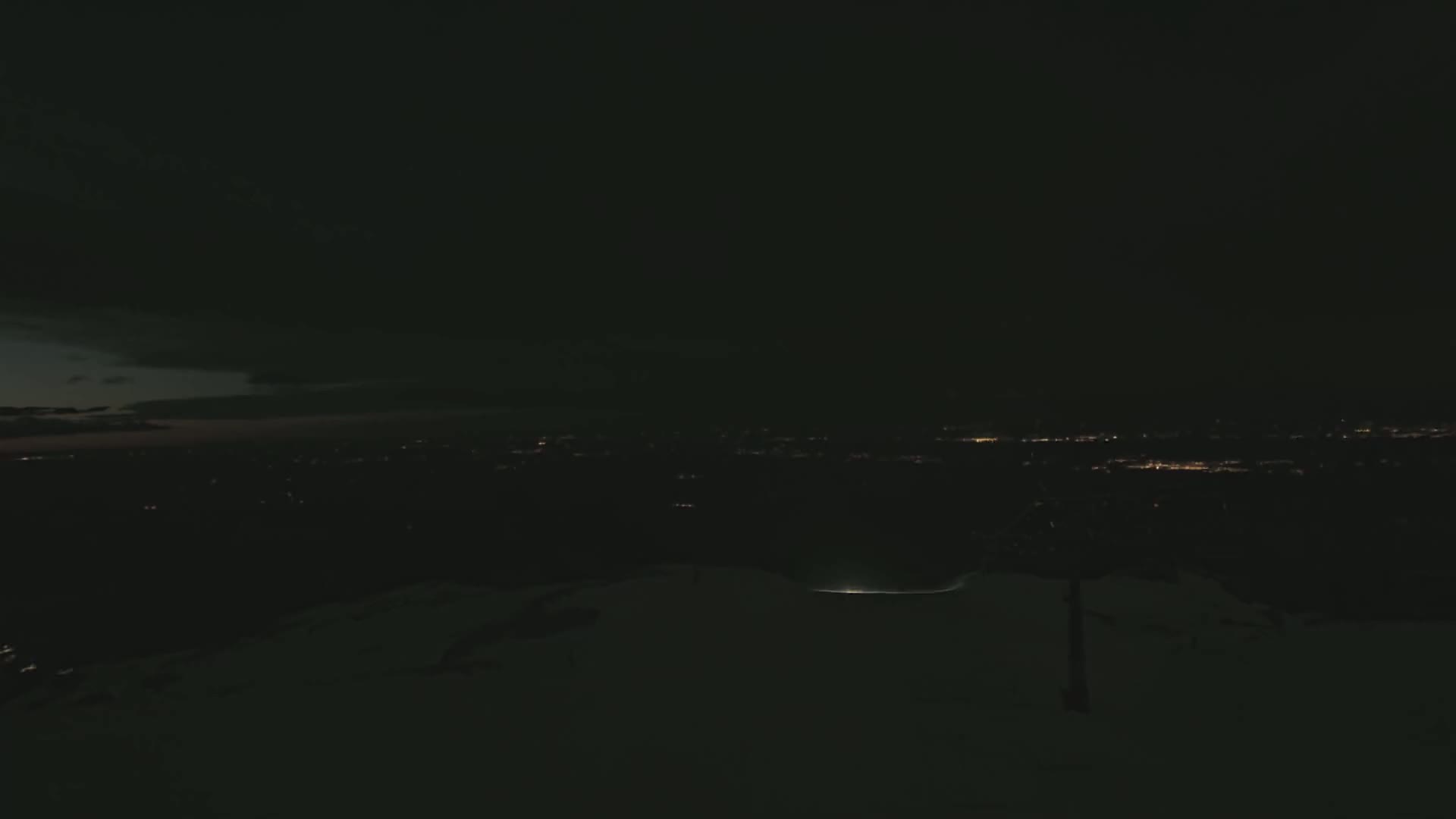 A dark, snowy landscape at night shows a distant city illuminated by faint lights under a mostly cloudy sky.